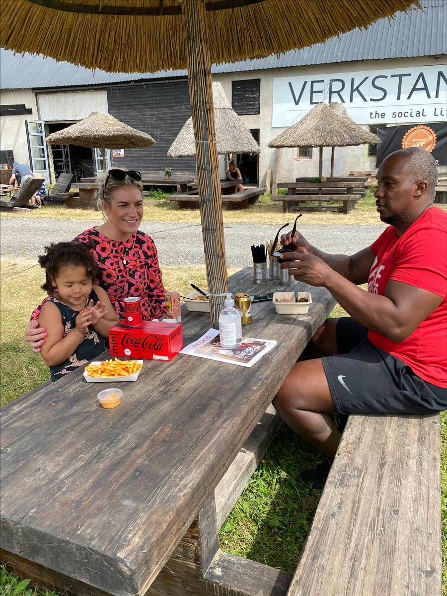 Gäster äter vid ett långbord i solen på Verkstan, med Joppes Food-skylten i bakgrunden